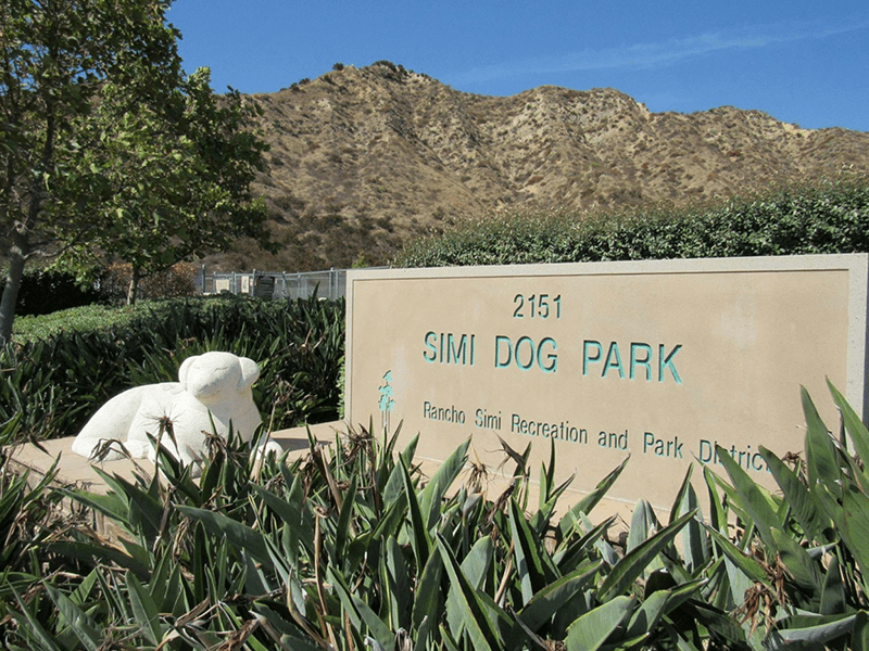 Simi Dog Park sign with plants and a dog statue in front. There are trees and hills in the background.