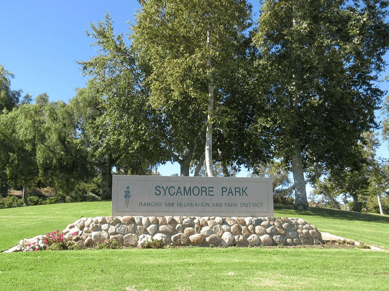 Sycamore Park sign with rocks and grass around it. Trees are in the background.