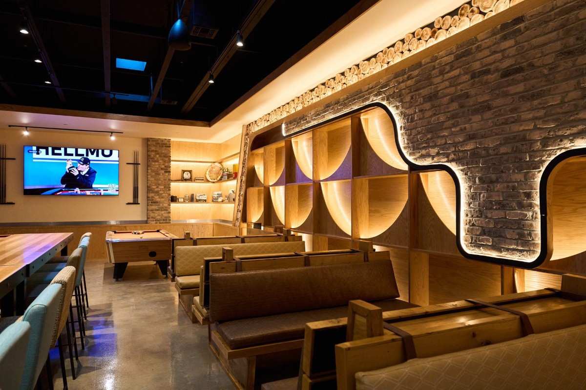 Modern lounge with wooden benches, a pool table, and a TV mounted on a brick wall; illuminated shelves and exposed ceiling create a contemporary atmosphere.
