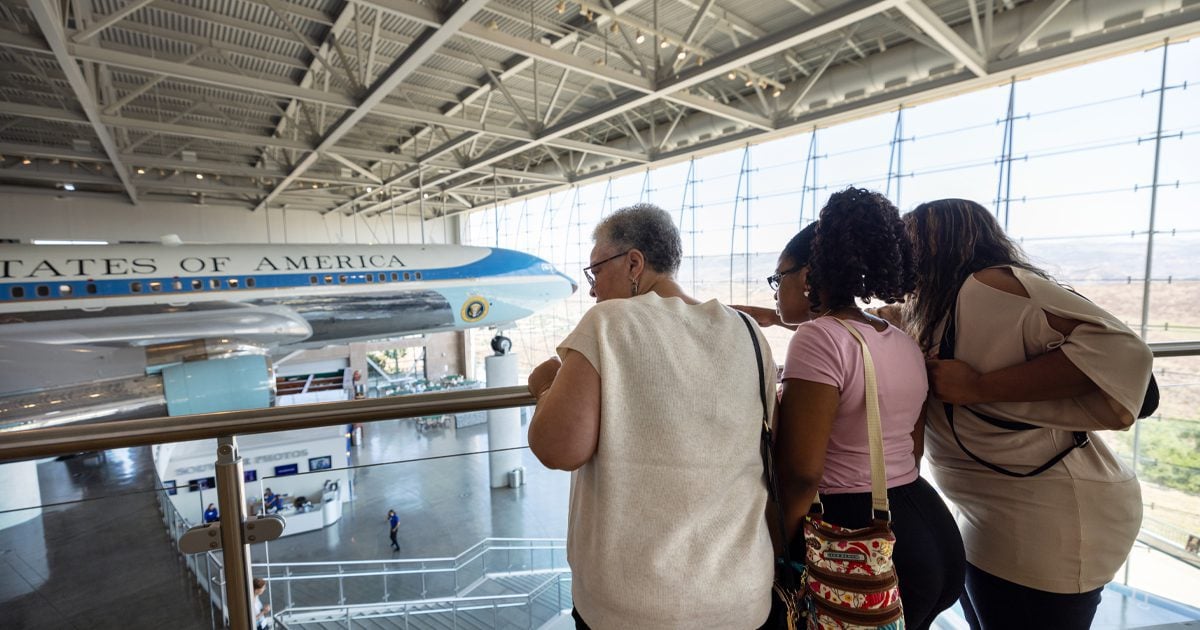 Three people stand on a balcony overlooking Air Force One displayed inside an aviation museum.