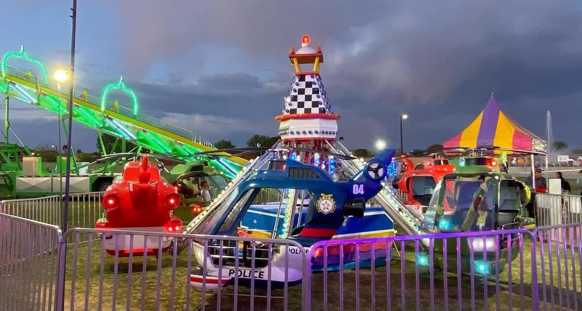 Small amusement park ride with police helicopter and fire truck cars, fenced in the foreground, with a slide and a striped tent in the background at dusk.