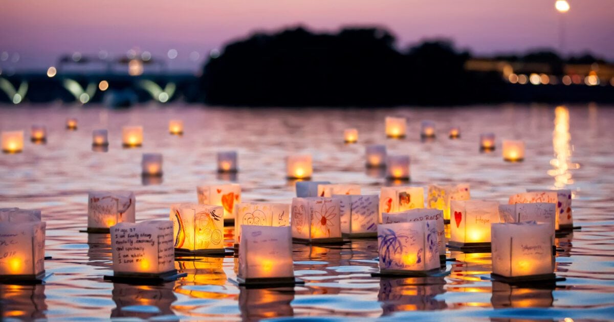 Paper lanterns with candles float on a calm body of water at sunset, with decorated messages visible on some lanterns and a blurred background of trees and lights.