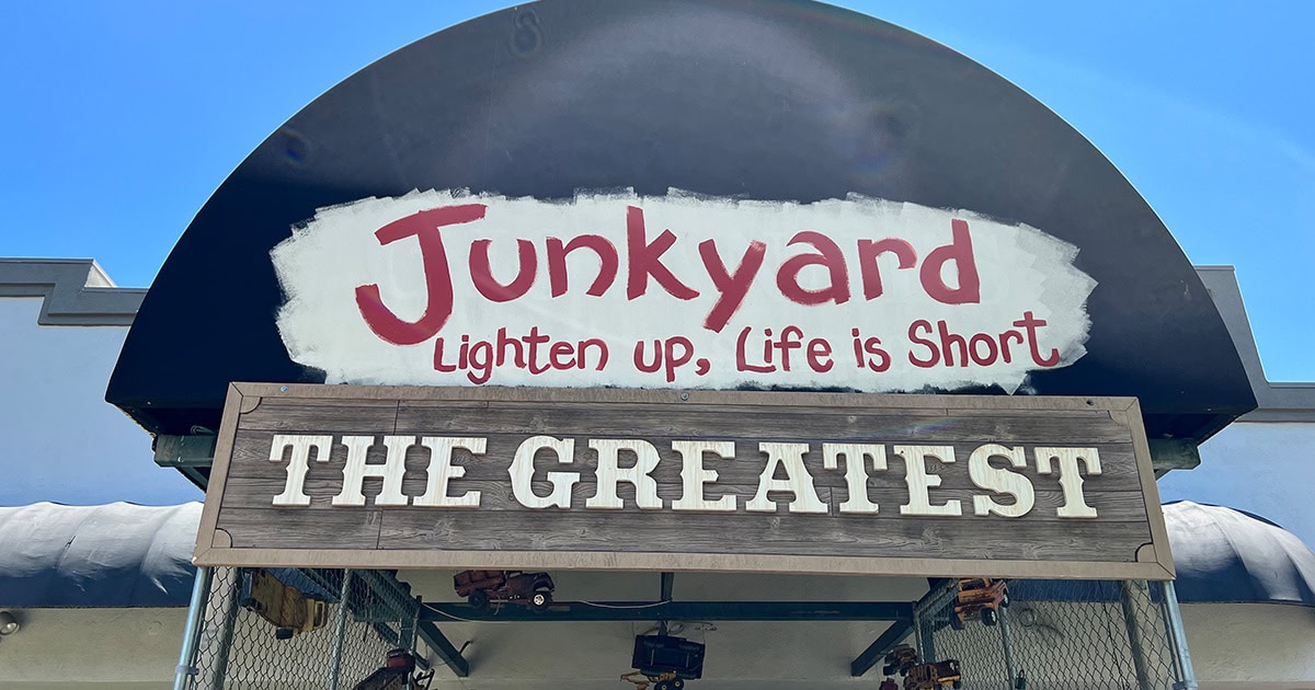 A sign reads “Junkyard: Lighten up, Life is Short” above a wooden sign with the words “THE GREATEST.” The signs are part of a building facade.